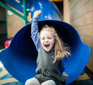 A little girl riding on top of a blue slide.